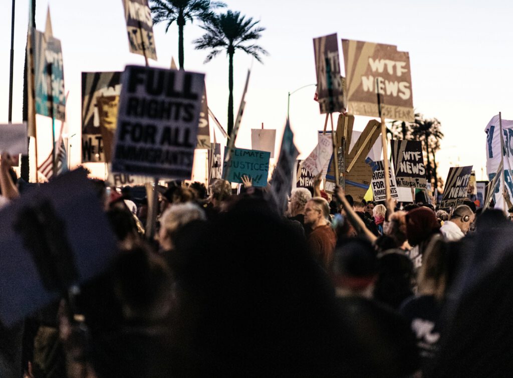 A diverse crowd gathers outdoors holding protest banners and signs at dusk.